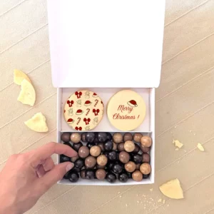 Coffret gourmand de Noël avec deux biscuits personnalisés “Merry Christmas” et assortiment de chocolats en boîte blanche à reliefs dorés.