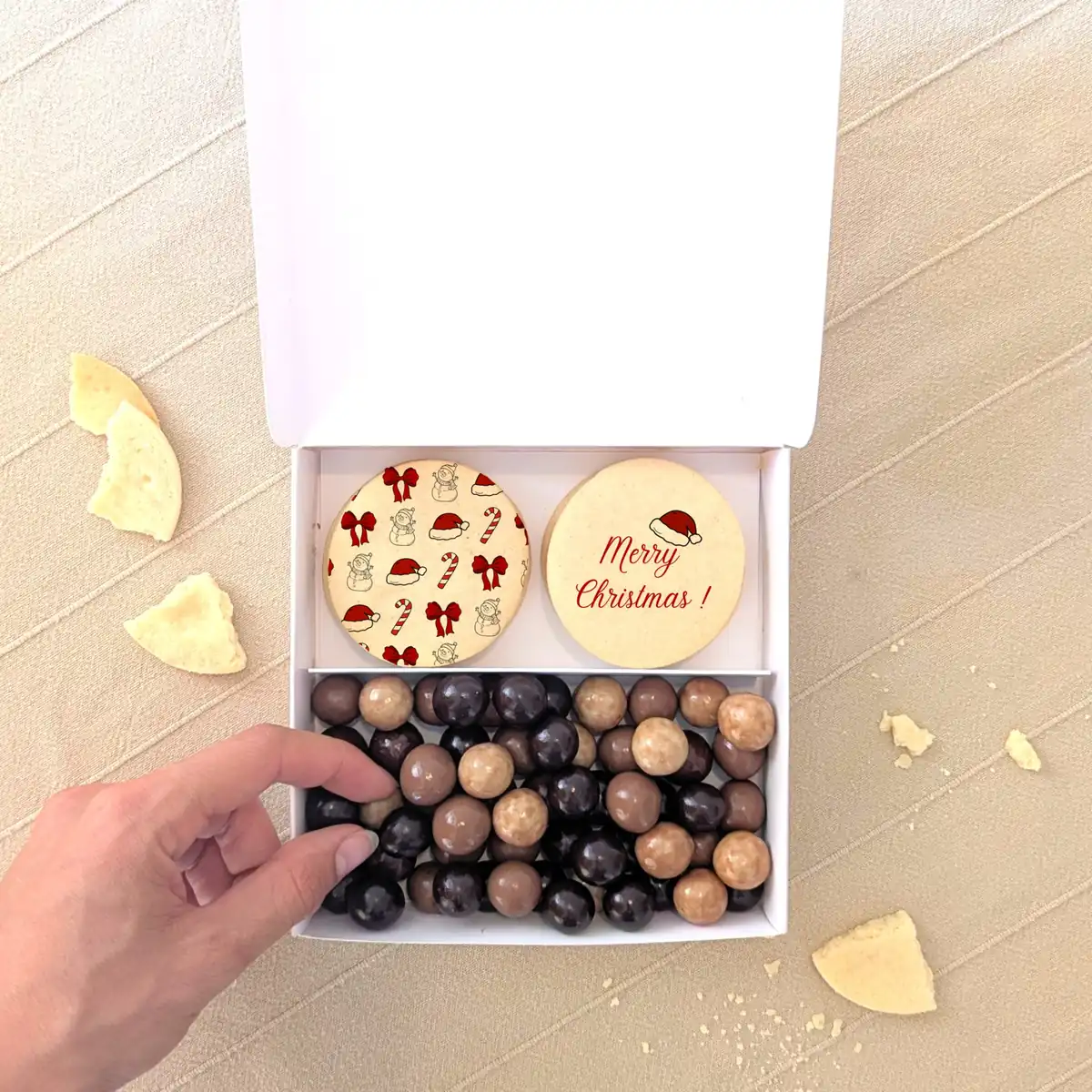 Coffret gourmand de Noël avec deux biscuits personnalisés “Merry Christmas” et assortiment de chocolats en boîte blanche à reliefs dorés.