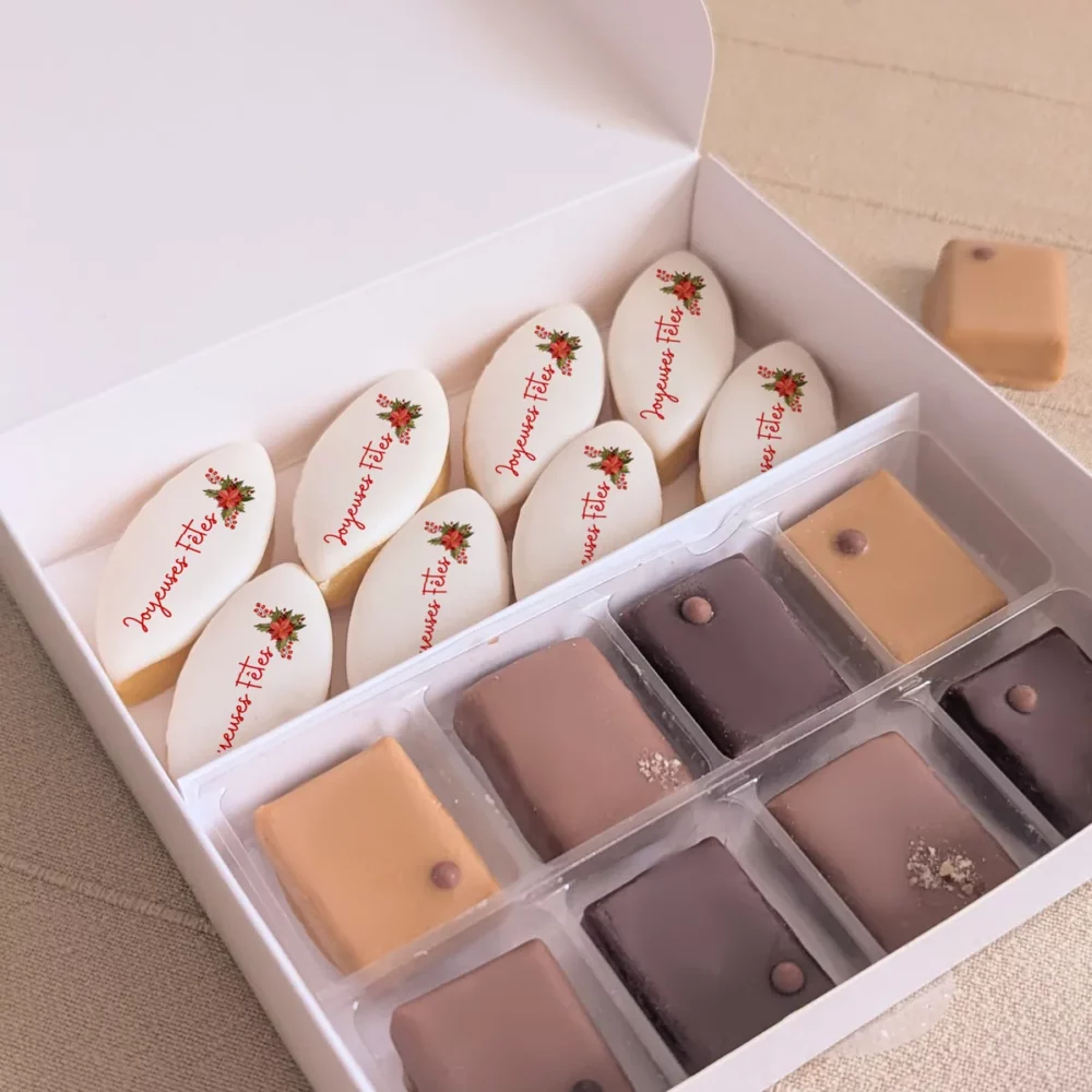 Gros plan sur les calissons personnalisés “Joyeuses Fêtes” et chocolats dans un coffret blanc aux reliefs dorés.
